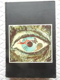 Examine a vintage book with a striking cover design featuring an eye, a figure in red, and surreal imagery.
