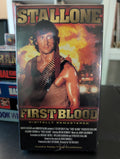 Sylvester Stallone stars in the action movie "First Blood," featuring Rambo with ammunition belt against a fiery background on the VHS cover.
