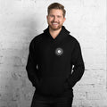 A smiling man models a black hoodie featuring a vinyl record label logo against a white brick wall.
