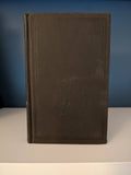 A worn, hardcover antique book stands upright on a white shelf against a blue wall.
