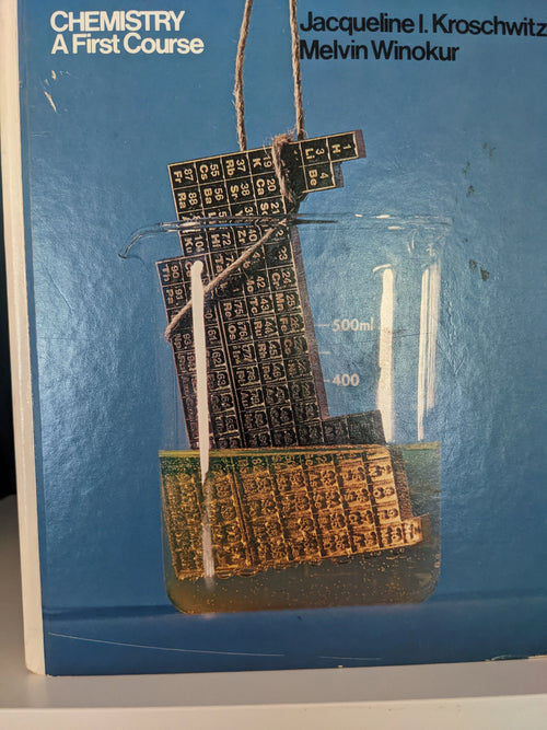 Chemistry textbook cover shows a periodic table partially submerged in a 500ml beaker.
